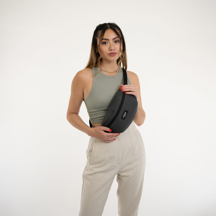 Frau trägt anthrazitgraue LARKSON Bauchtasche Finn über hellem Top und beigen Hosen