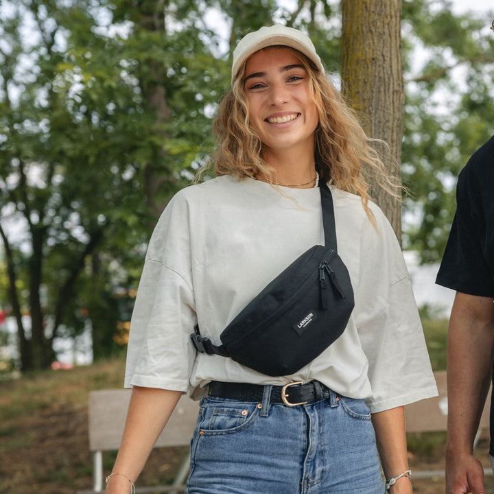 Frau im Freien trägt beige T-Shirt, Jeans und schwarze Bauchtasche 'Finn' von Larkson quer über der Brust