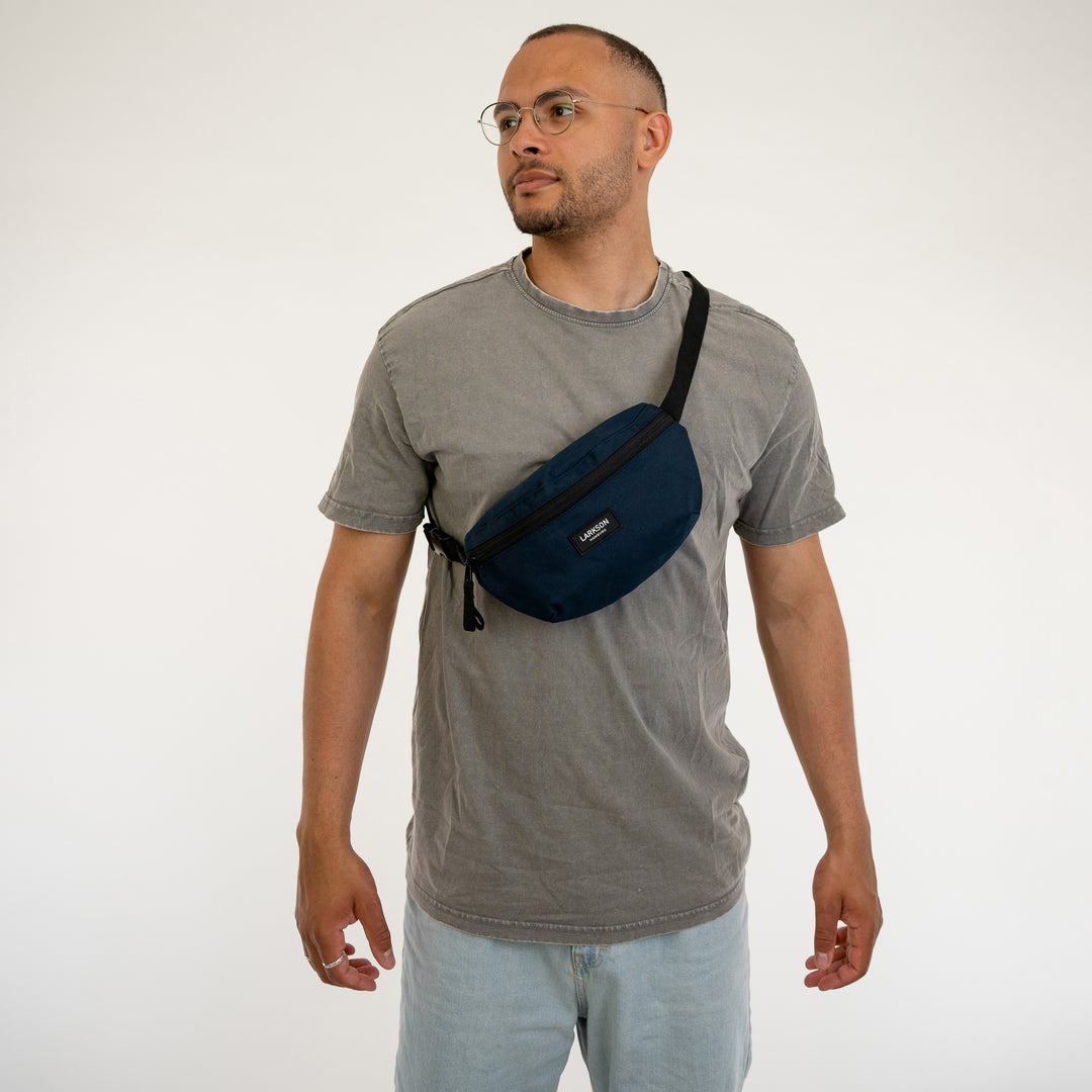 Mann trägt dunkelblaue LARKSON Bauchtasche Finn quer über den Oberkörper mit grauem T-Shirt und hellen Jeans