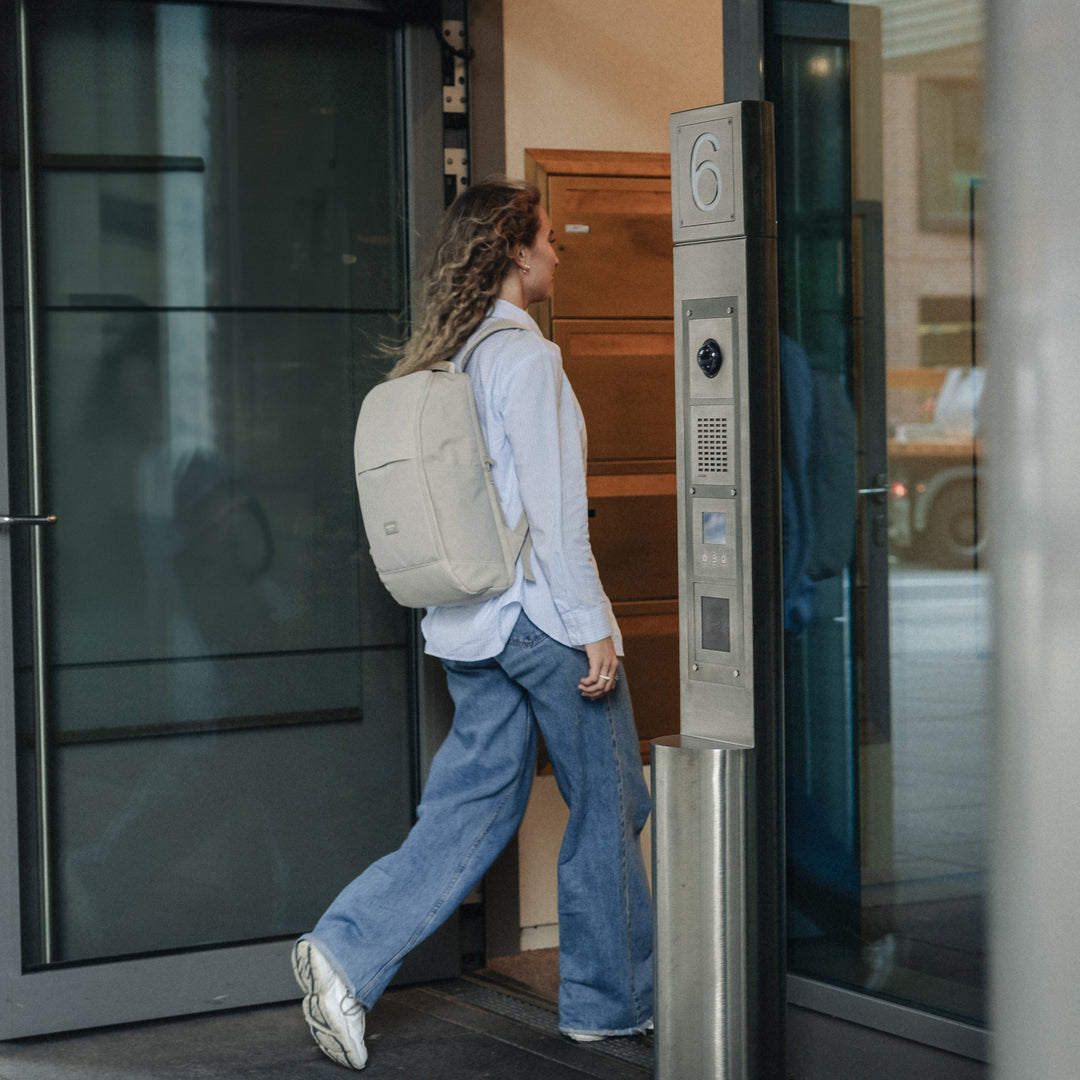 Frau mit beigem Rucksack Niklas in Business-Design trägt weiße Bluse und blaue Jeans und betritt ein Gebäude