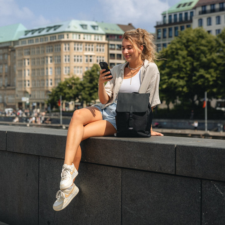 Frau sitzt auf einer Mauer in der Stadt mit schwarzem Damenrucksack 'Ida' von Larkson, trägt Shorts und weiße Schuhe.