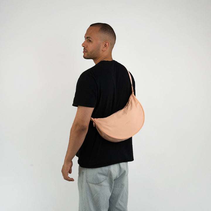 Mann mit schwarzem T-Shirt trägt pfirsichfarbenen Half Moon Bag Solveig Medium quer am Rücken