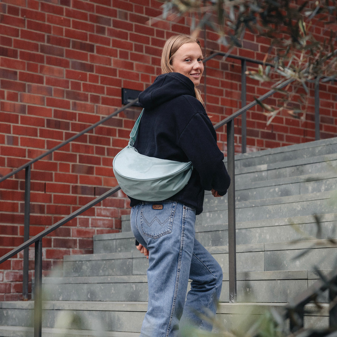 Frau trägt mittlere Half Moon Bag Solveig in Mint Farbe über der Schulter vor einer Treppe mit roter Backsteinmauer