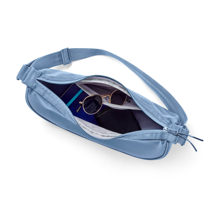 Hellblaue Half Moon Bag Solveig Medium mit verstellbarem Träger, offen und mit Sonnenbrille, Buch und Geldbörse gefüllt