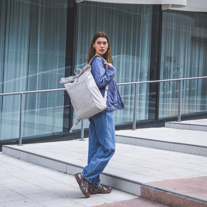 Frau mit beigem Rolltop Rucksack No 2 von LARKSON, Jeans und blaue Jacke, städtische Umgebung