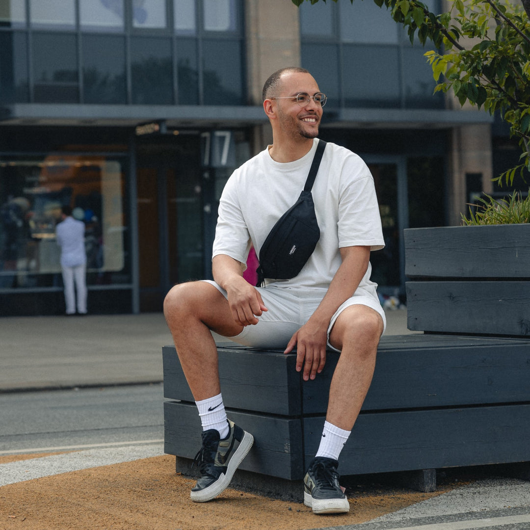 Mann trägt schwarze LARKSON Bauchtasche Ole über weißem T-Shirt und Shorts, sitzt auf Bank
