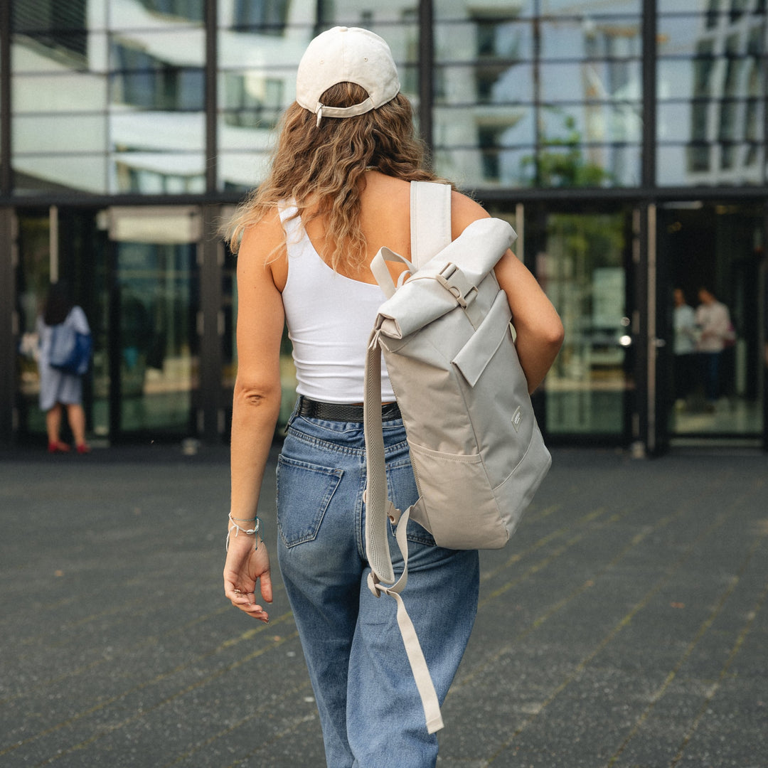 Frau von hinten mit beige Rolltop Rucksack No 4 von LARKSON, weißem Tanktop, Jeans und hellbeiger Kappe vor modernem Gebäude