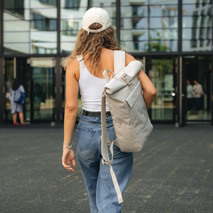 Frau von hinten mit beige Rolltop Rucksack No 4 von LARKSON, weißem Tanktop, Jeans und hellbeiger Kappe vor modernem Gebäude