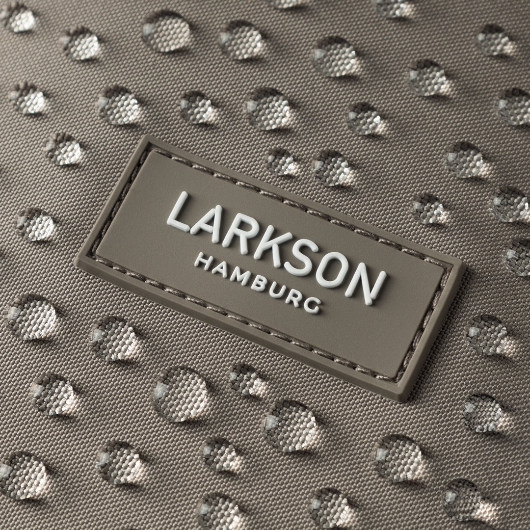 Nahaufnahme des LARKSON Markenlabels auf einem wasserabweisenden grauen Rolltop Rucksackstoff mit Wassertropfen