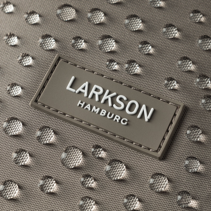 Nahaufnahme des LARKSON Markenlabels auf einem wasserabweisenden grauen Rolltop Rucksackstoff mit Wassertropfen