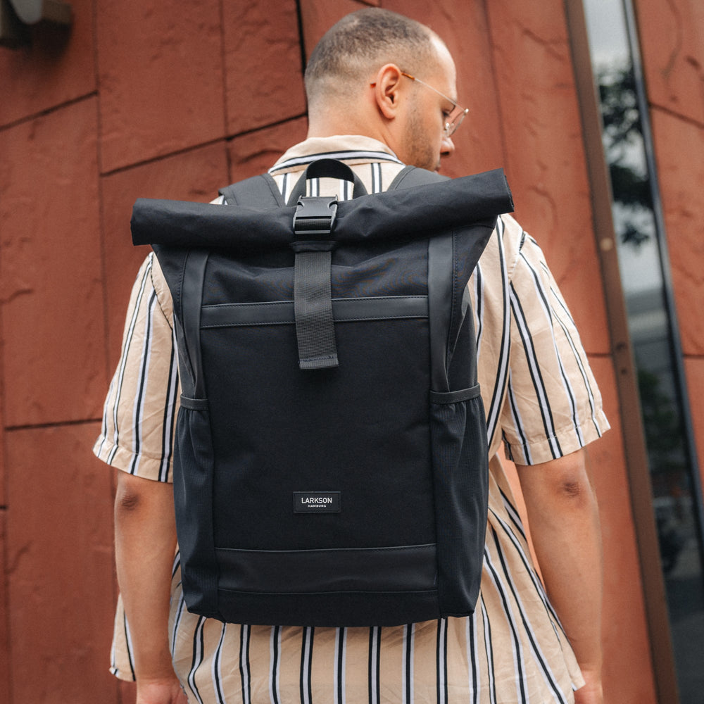 Mann trägt schwarzen LARKSON Rolltop Rucksack No 2 vor roten Wand