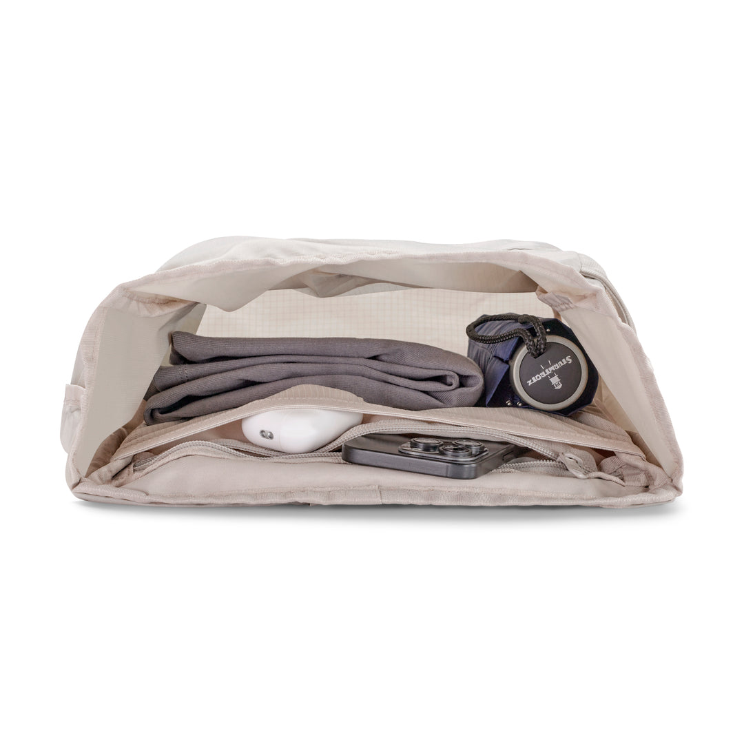 Beiger faltbarer Rolltop Rucksack Arne liegend mit Kamera, zusammengefalteter Kleidung und Elektronik