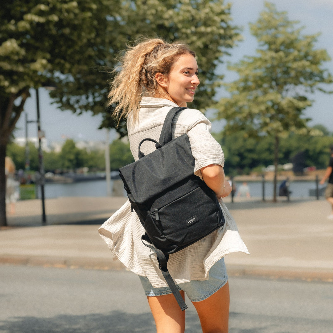 Frau mit blondem Pferdeschwanz trägt schwarzen LARKSON Rucksack Ronja, trägt helle Bluse und Jeansshorts im Freien
