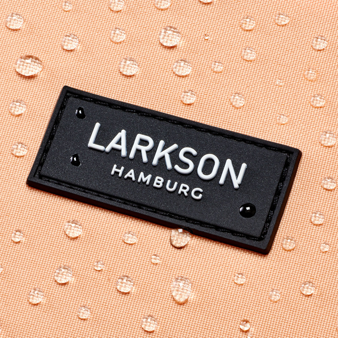 Nahaufnahme des LARKSON Hamburg Logos auf einem pfirsichfarbenen, wasserabweisenden Schulrucksackstoff mit Wassertropfen