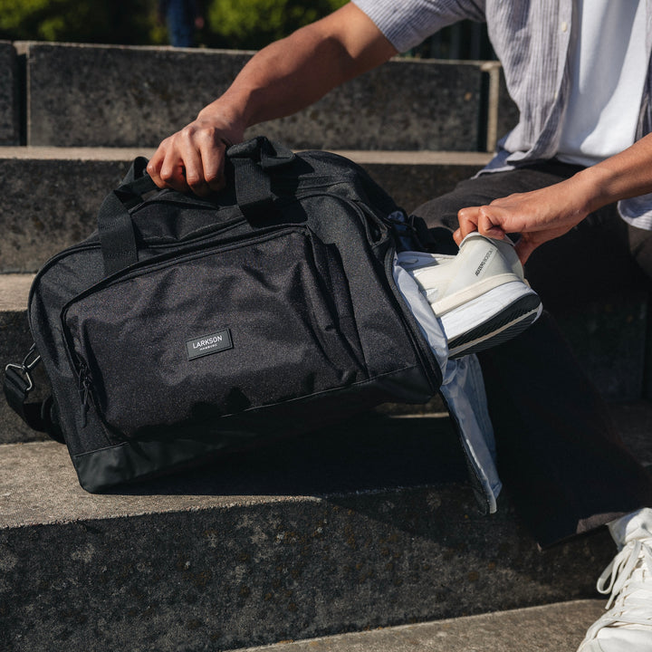 Schwarze LARKSON Reisetasche Finja mit Reißverschlussfach, eine Hand hält weißen Sportschuh