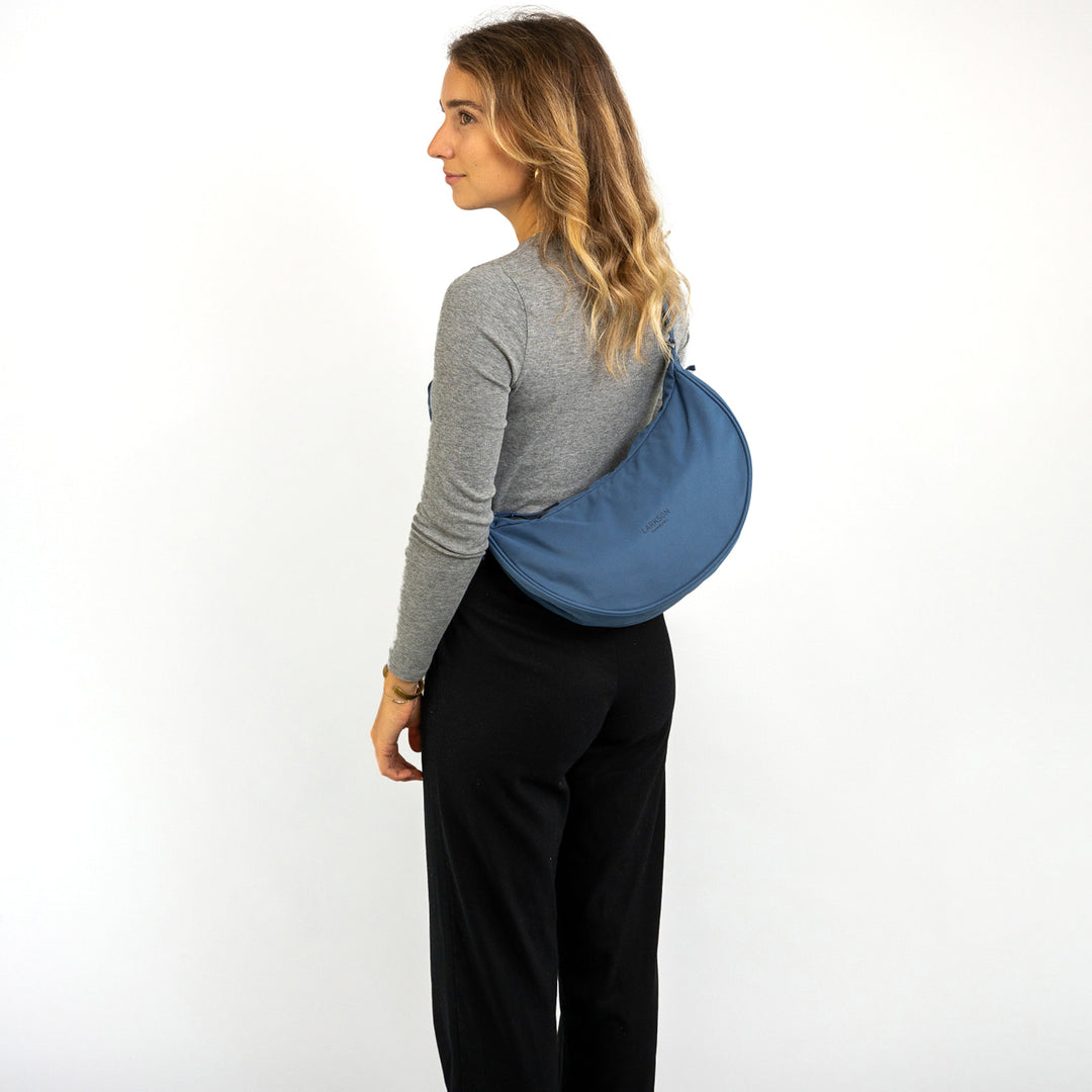 Frau trägt blaue Half Moon Bag Solveig Medium als Umhängetasche, grauer Langarmpullover, schwarze Hose