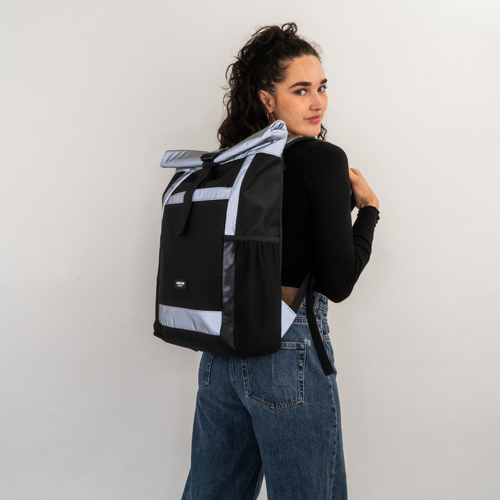 Frau mit schwarzem und silber-reflektierendem Rolltop Rucksack No 2 von LARKSON trägt schwarze Oberteile und blaue Jeans