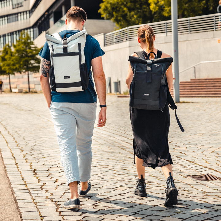 Zwei Personen tragen Rolltop Rucksack No 2 Rucksäcke von LARKSON, eine in weiß, eine in schwarz, urbaner Hintergrund