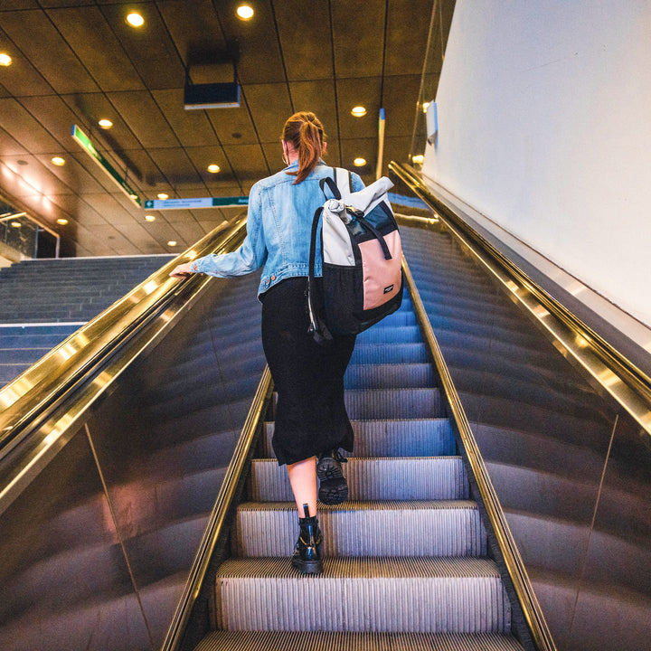 Frau trägt Rolltop Rucksack No 2 in Grau-Rosa, steigt Rolltreppe in urbanem Gebäude hinauf