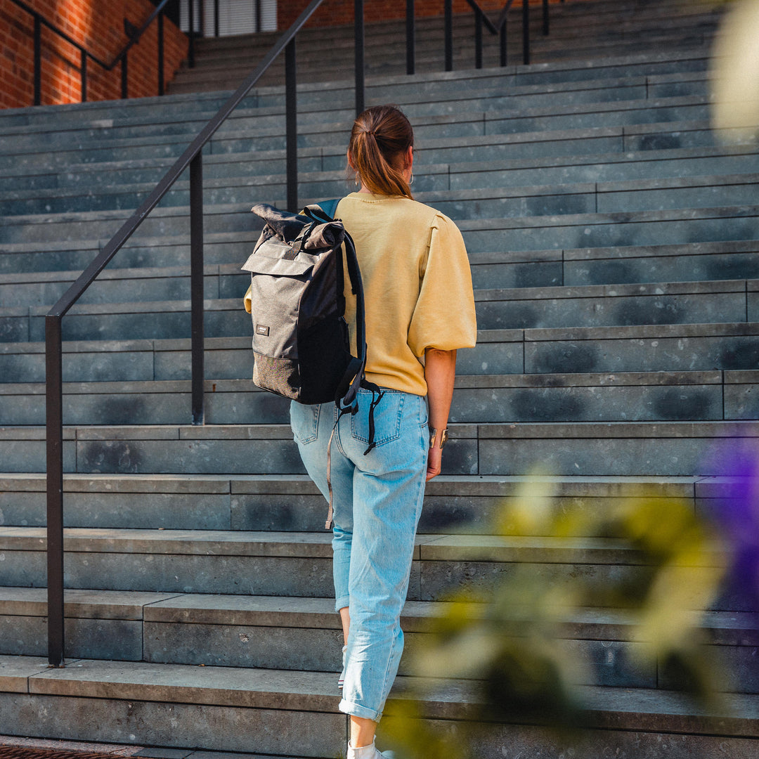 Rucksack Rolltop Rucksack No 4 in Schwarz aus Stoff an einer Frau mit gelbem Oberteil und blauer Jeans auf einer Treppe