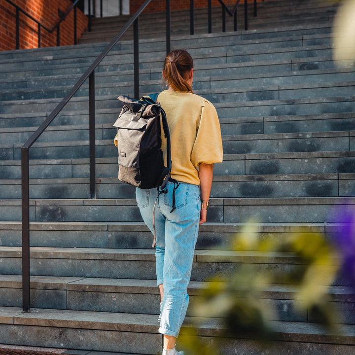 Rucksack Rolltop Rucksack No 4 in Schwarz aus Stoff an einer Frau mit gelbem Oberteil und blauer Jeans auf einer Treppe