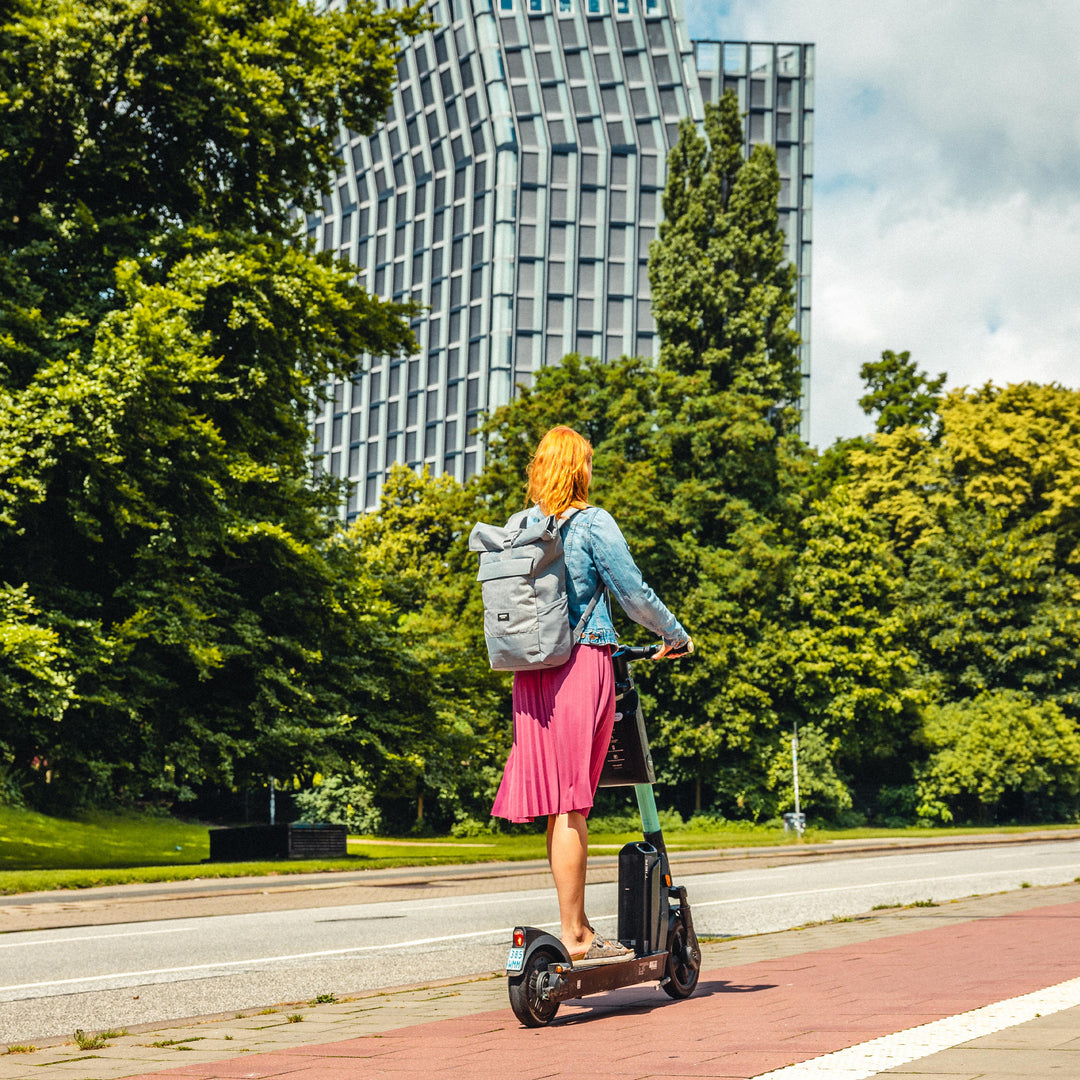 Frau mit hellgrauem Rolltop Rucksack No 4 von LARKSON auf einem E-Scooter vor urbaner Kulisse mit Bäumen und Gebäude