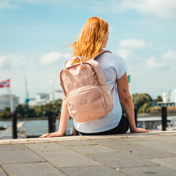 Frau mit rosa LARKSON Rucksack No 6 sitzt an Wasser mit Blick auf Stadt und Himmel