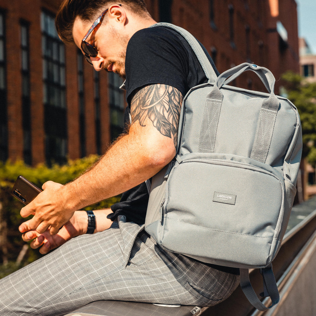 Mann mit Sonnenbrille sitzt draußen und trägt grauen LARKSON Rucksack No 6 auf dem Rücken
