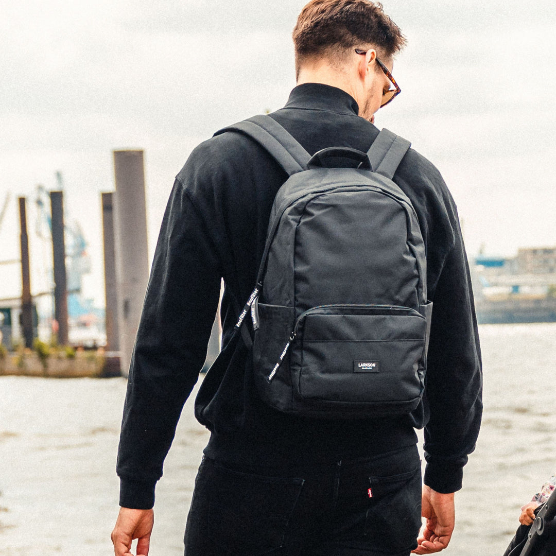 Junger Mann trägt schwarzen Rucksack No 3 von LARKSON, stehend am Wasser