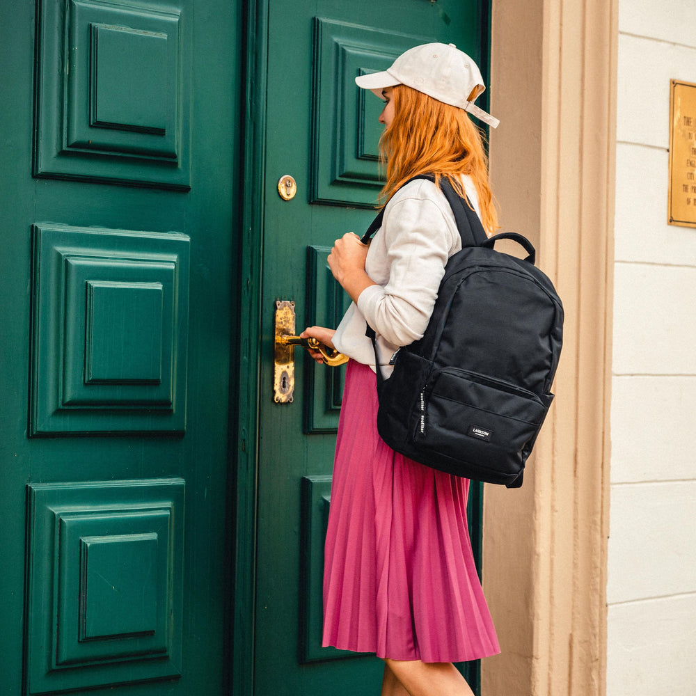 Teenagermädchen mit weißem Basecap und pinkem Rock trägt LARKSON schwarzen Rucksack und öffnet grüne Tür