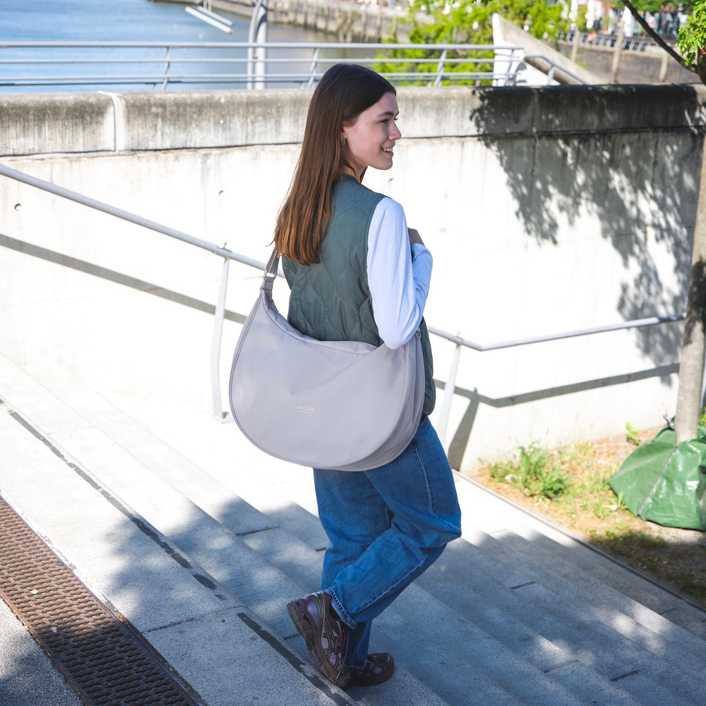 Frau mit halbmondförmiger, beiger XL-Tasche Half Moon Bag Solveig von LARKSON über der Schulter, trägt grüne Weste und blaue Jeans