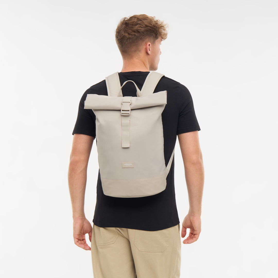 Mann trägt hellbeigen Rolltop Rucksack Janne Small von LARKSON, schwarze T-Shirt, beige Hose