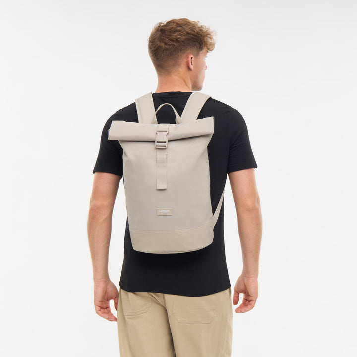Mann trägt hellbeigen Rolltop Rucksack Janne Small von LARKSON, schwarze T-Shirt, beige Hose