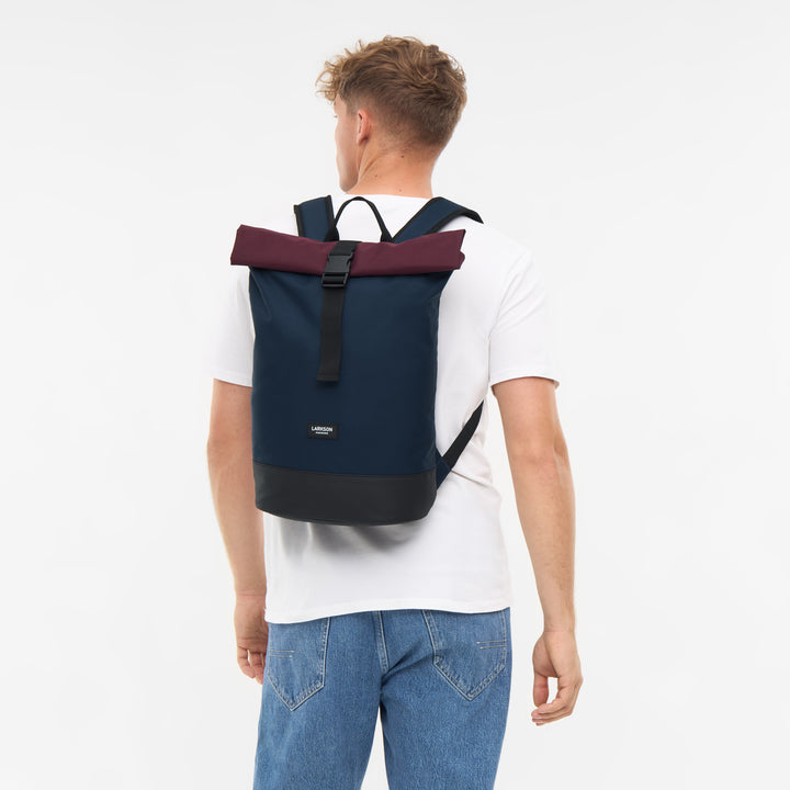 Mann trägt blauen LARKSON Rolltop Rucksack Janne Small mit bordeauxroter Oberseite und schwarzem Boden