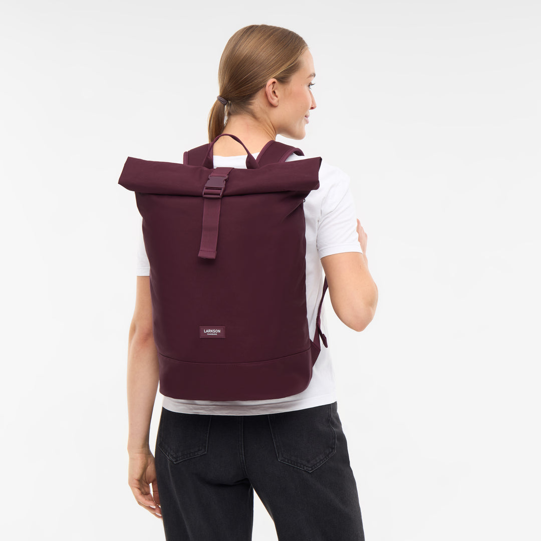 Frau trägt bordeauxfarbenen Rolltop Rucksack Janne von LARKSON, weißes T-Shirt, schwarze Hose