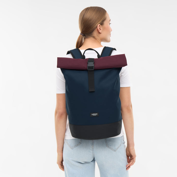 Rolltop Rucksack Janne von LARKSON in marineblau mit burgunderrotem Rollverschluss getragen von Frau mit weißem T-Shirt