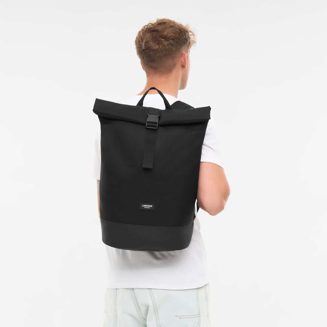 Herren mit weißem T-Shirt trägt schwarzen Rolltop Rucksack Janne von Larkson mit Logo vorne