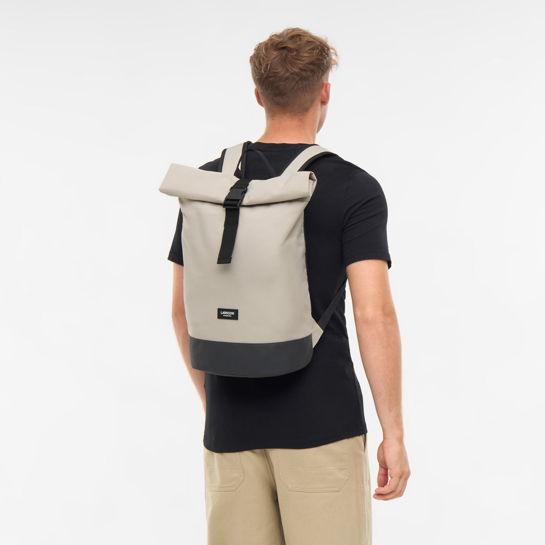 Beiger Rolltop-Rucksack Janne von LARKSON mit schwarzem Boden, getragen von einem Mann in schwarzem T-Shirt und beigen Hosen