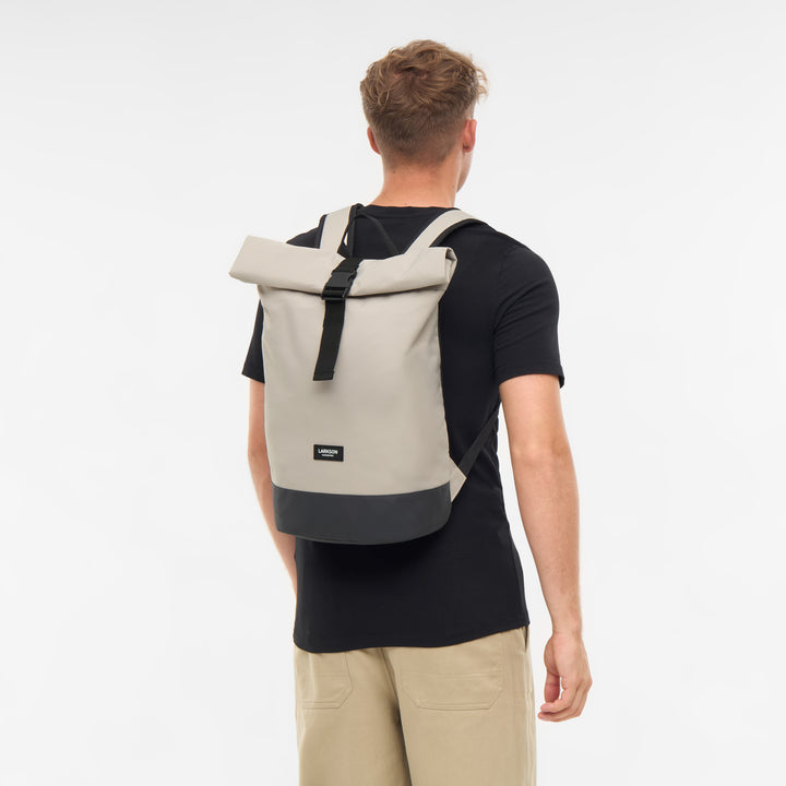 Beiger Rolltop-Rucksack Janne von LARKSON mit schwarzem Boden, getragen von einem Mann in schwarzem T-Shirt und beigen Hosen