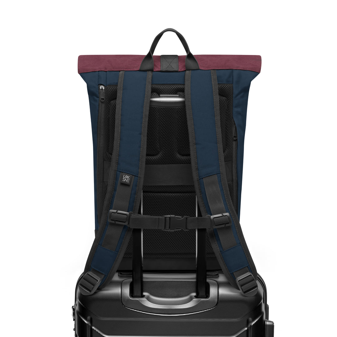 Rolltop-Rucksack Janne Small in Blau mit bordeauxrotem Rand von hinten, auf einem Trolleygriff befestigt