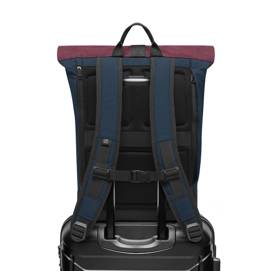 Rückansicht des Rolltop Rucksacks Janne in Blau mit Beerenton am oberen Rand, auf einem schwarzen Koffer stehend