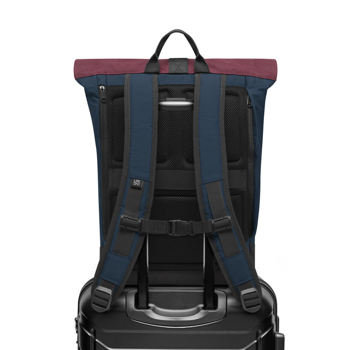 Rückansicht des Rolltop Rucksacks Janne in Blau mit Beerenton am oberen Rand, auf einem schwarzen Koffer stehend