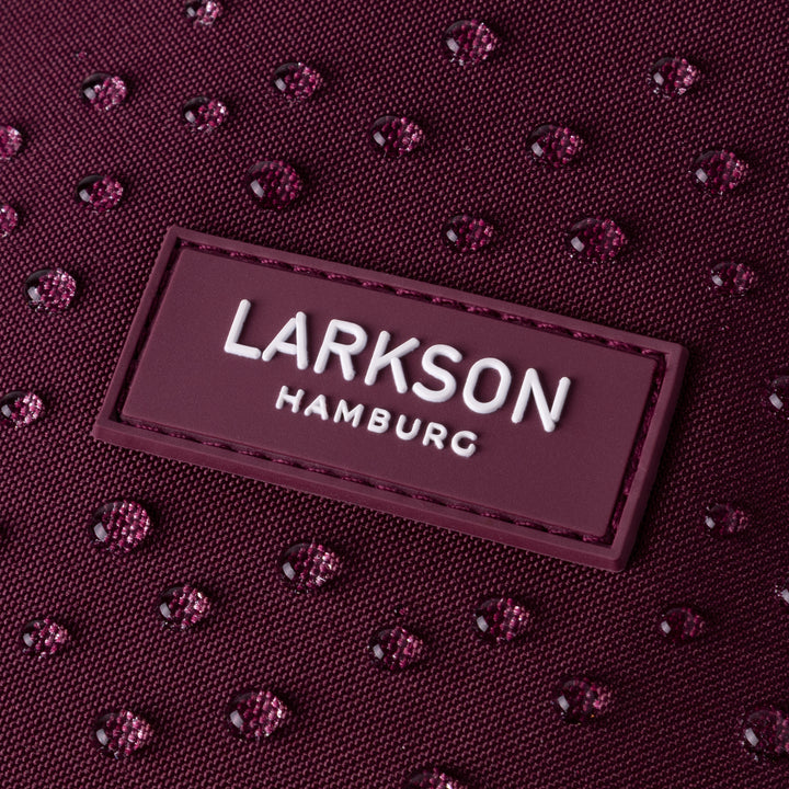 Nahaufnahme des LARKSON Hamburg Logos auf einem wasserabweisenden, dunkelroten Rolltop Rucksack Janne mit Wassertropfen